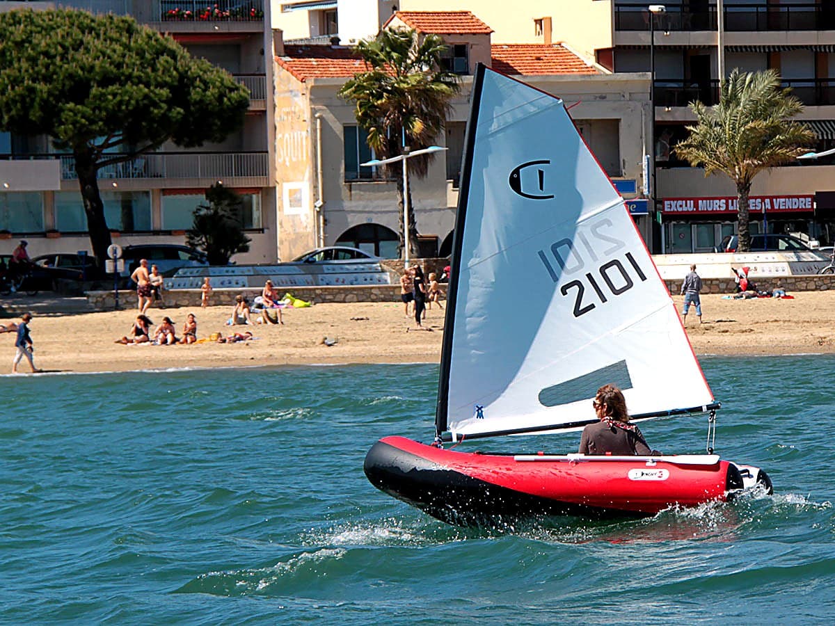 The original DinghyGo sailing past a Côte d'Azur beach in 2013 — red-and-white inflatable hull with a small sail, palm trees and pastel-stuccoed seafront buildings behind, sunbathers on the sand